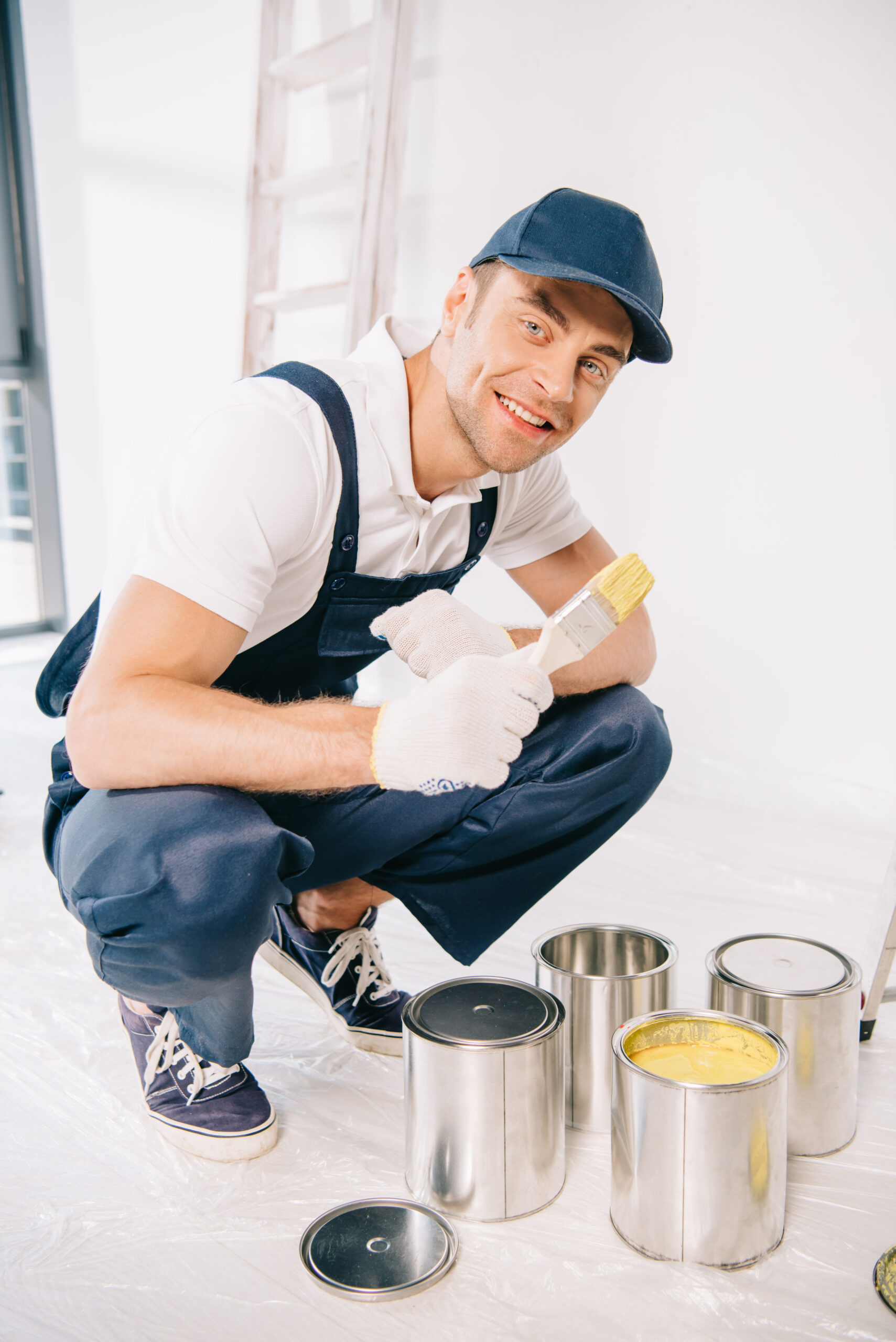 handsome young painter in uniform holding paintbrush near cans with paint and smiling at camera Reliaco Painting Fishers, IN